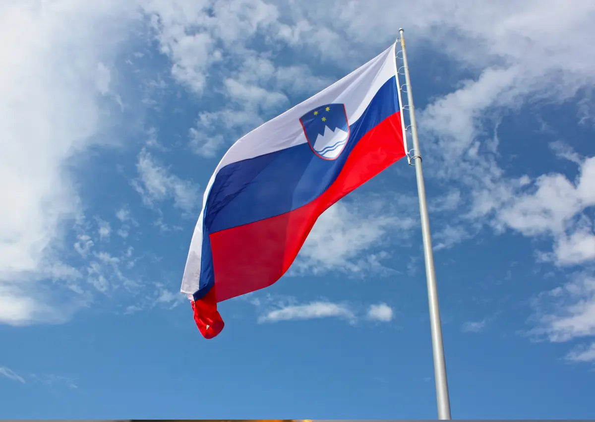 Die Nationalflagge von Slowenien mit den weiß-blau-roten Streifen und dem offiziellen Wappen weht im Wind vor blauem Himmel – Zur digitalen Vignette Slowenien (E-Vinjeta) auf mautbox24.com.